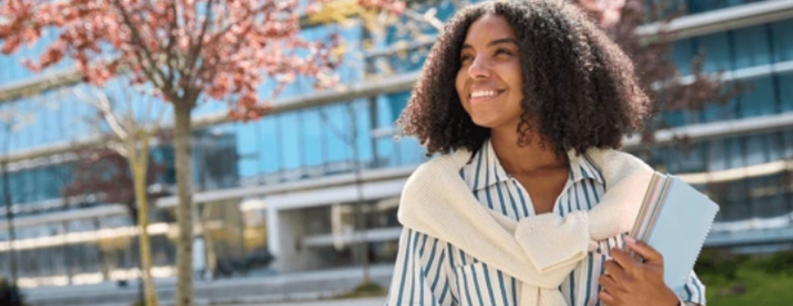 smiling young minority woman carrying books in front of a building
